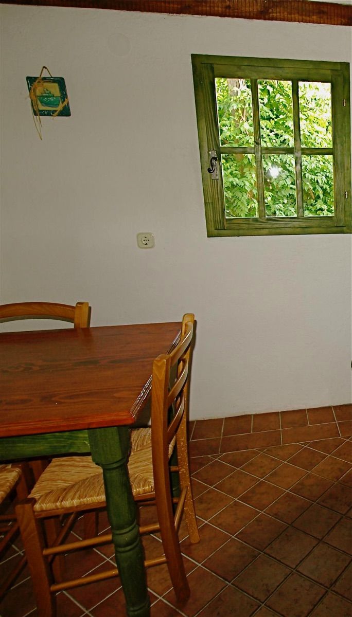 A dining area with a wooden table and chairs, a window with a green frame, and a wall-mounted electrical outlet.