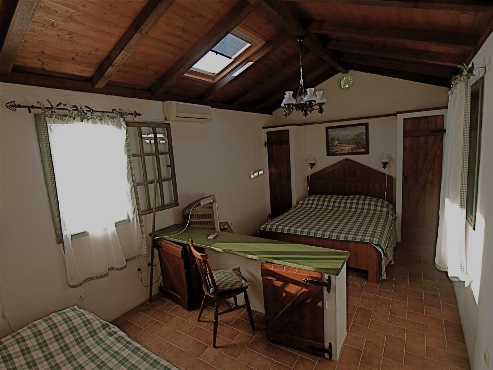 Een slaapkamer met een houten plafond, dakraam, een bed met een geruit laken, een bureau met stoel en een klein raam met gordijnen.