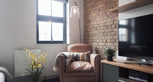 A modern living room with a brick wall, a leather armchair, a large flat-screen TV, and a potted plant.