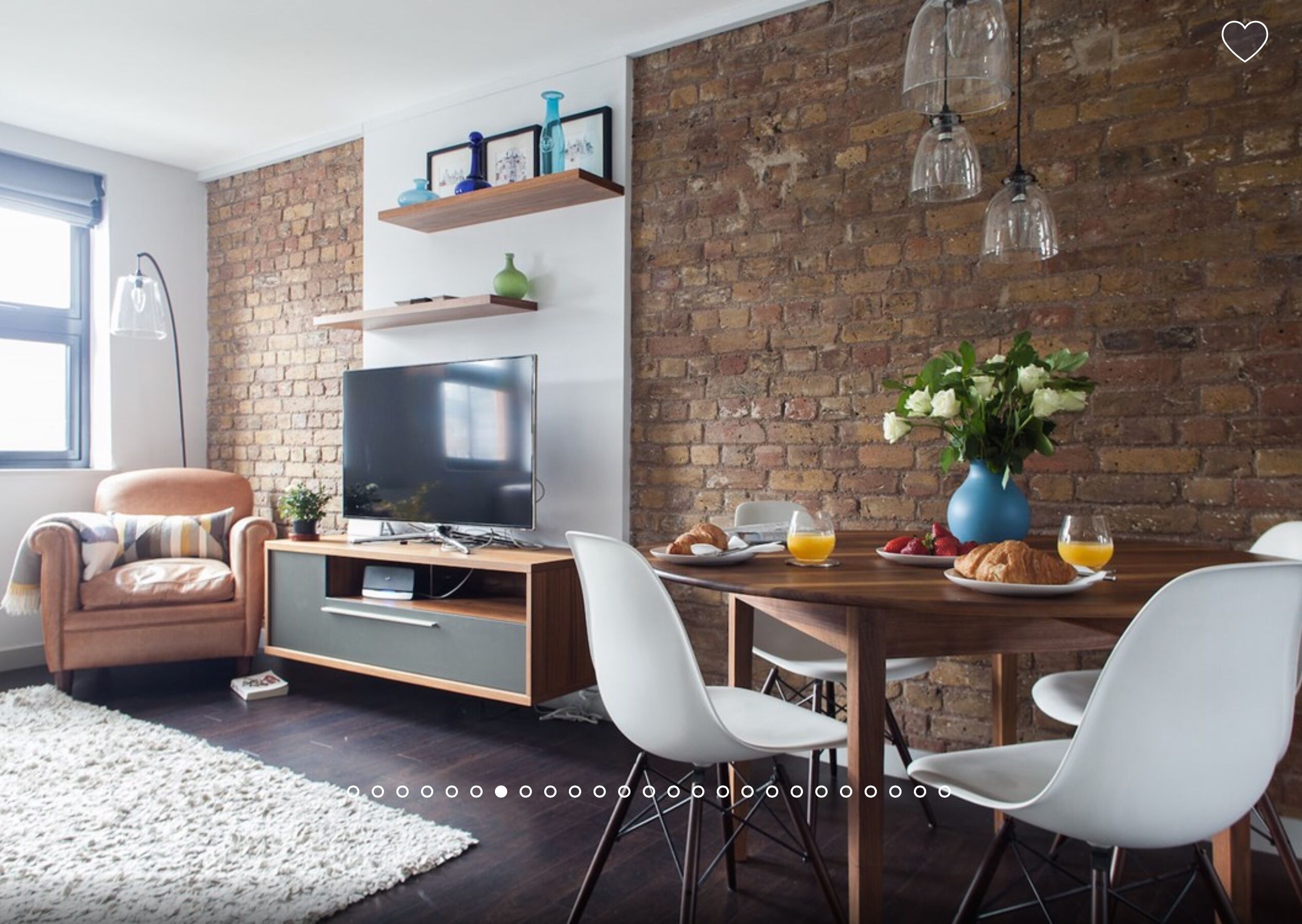 A modern living room with a brick wall, a television, a dining table with chairs, and a coffee table with breakfast items.