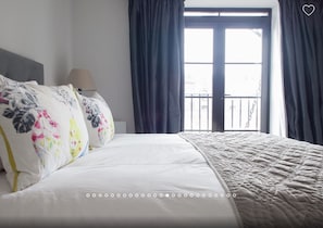 A hotel room with a bed, a nightstand, a lamp, and a view of the outside through a window with curtains.