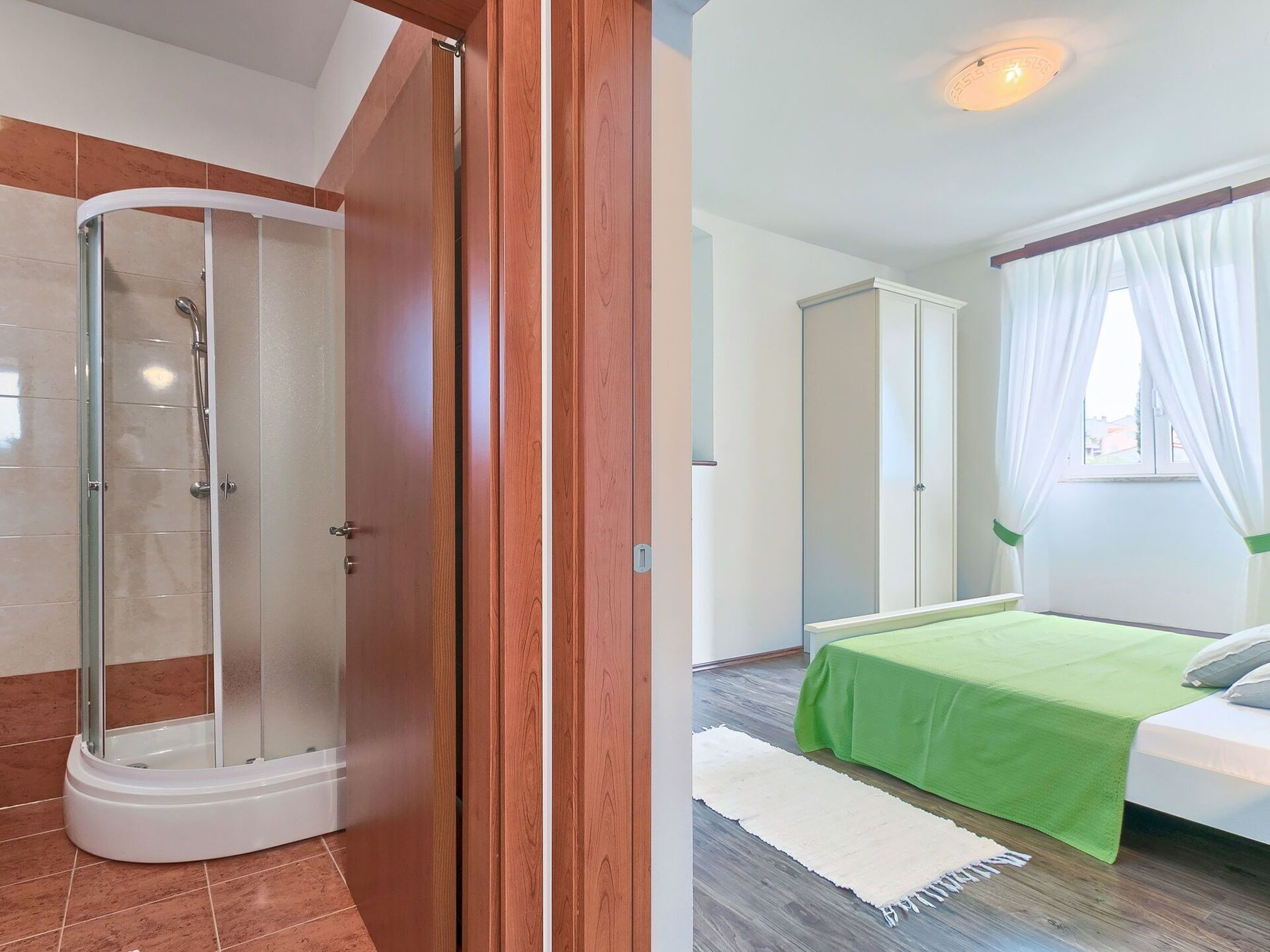 A bathroom with a glass shower enclosure and a bedroom with a bed and a window.