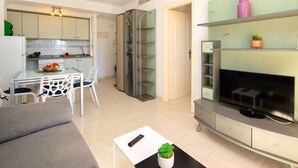 Un salon moderne avec un canapé gris, une table basse agrémentée d’une plante et un téléviseur posé sur un meuble. On aperçoit une cuisine avec un coin repas à l’arrière-plan.