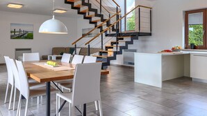 Eine moderne Küche mit einer zentralen Kochinsel, einem Essbereich mit Holztisch und weißen Stühlen sowie einer Treppe mit Holz- und schwarzen Geländern.