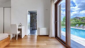 Ein modernes Hotelzimmer mit Holzboden, einem Schreibtisch mit Stuhl und einem großen Fenster mit Blick auf einen Pool und Grünflächen.