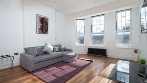 Ein modernes Wohnzimmer mit einem grauen Ecksofa, einem Glastisch und großen Fenstern mit Blick auf die Stadt.