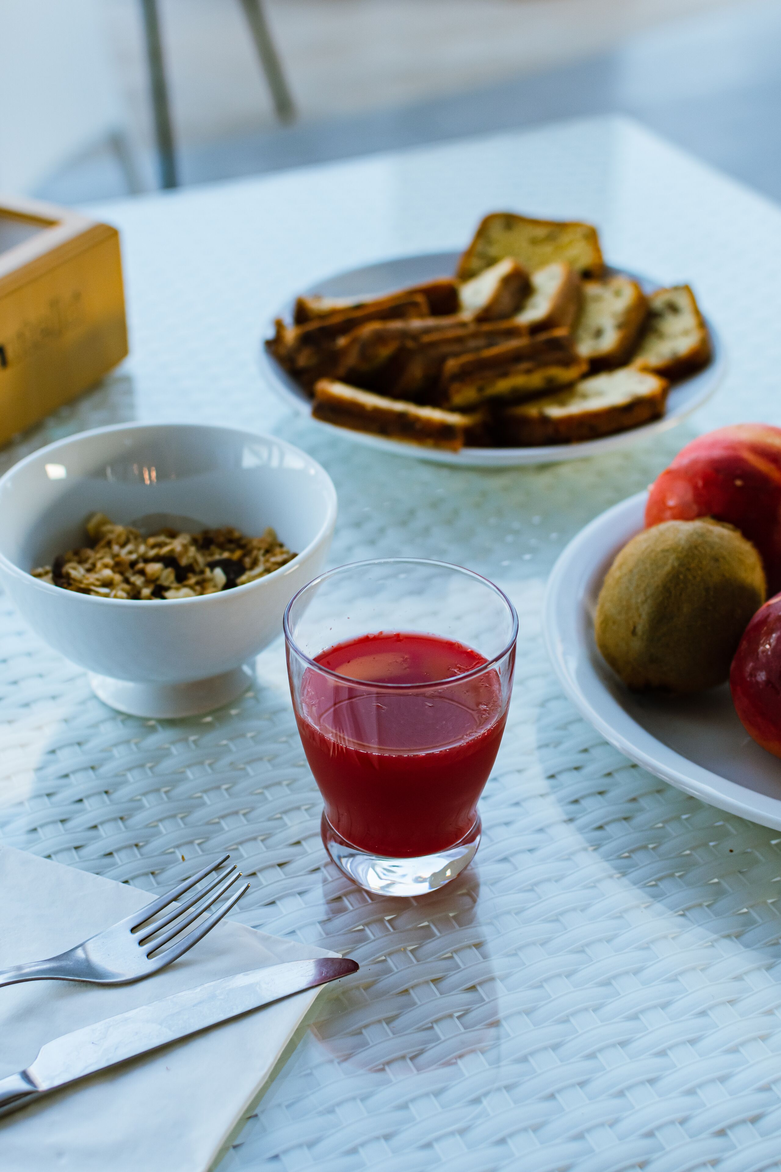 Un desayuno con tostadas, un tazón de cereal, un vaso de jugo de frutos rojos y un plato de kiwis sobre una mesa blanca.