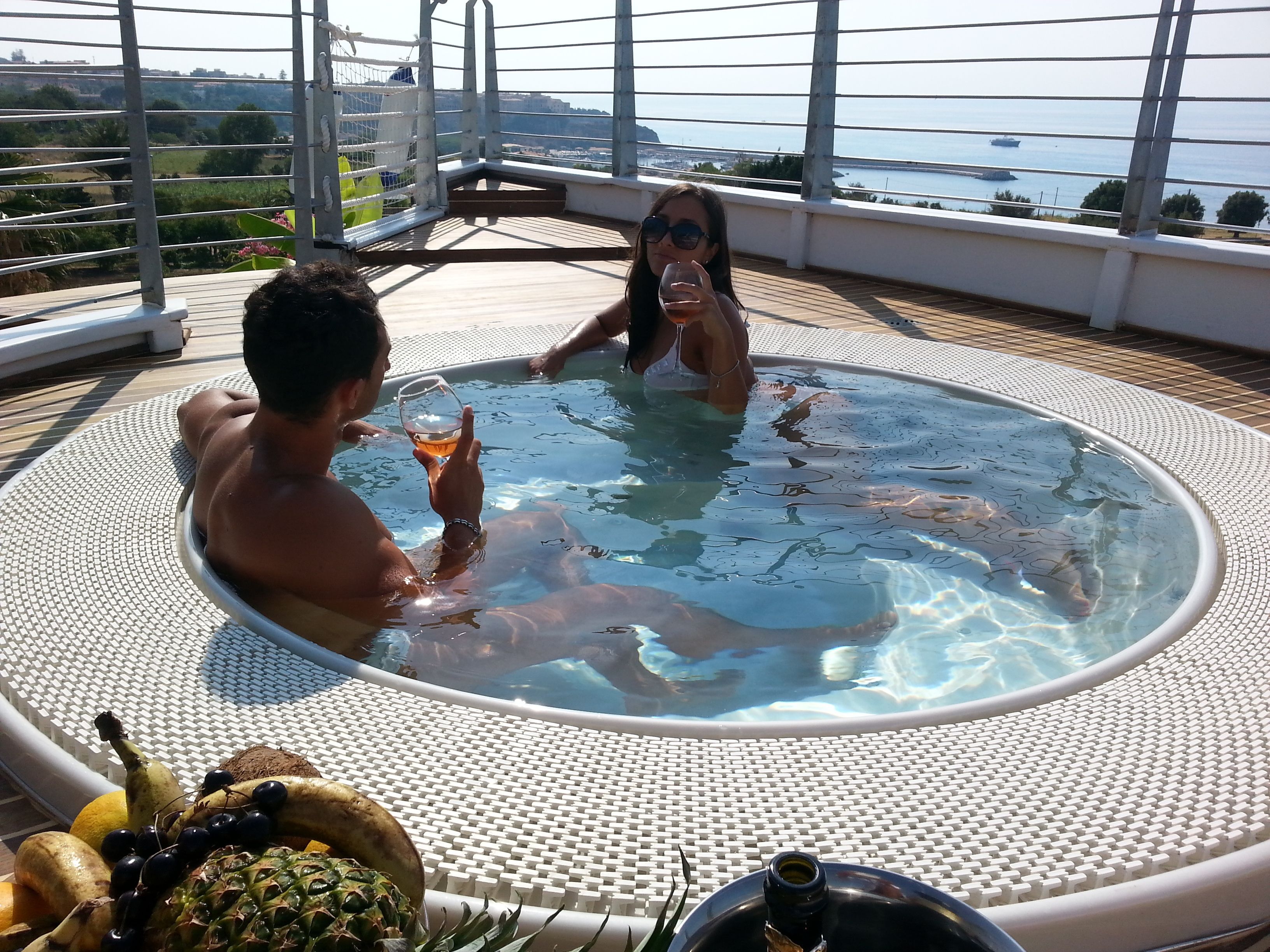 A hot tub with two people, surrounded by a deck with a view of the sea and boats.