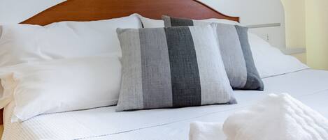 A neatly made bed with white linens and striped pillows, a wooden headboard, and a framed painting above it.