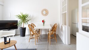 Una sala de estar moderna con una mesa blanca, cuatro sillas de mimbre, una planta en maceta, un televisor y un ventanal grande.