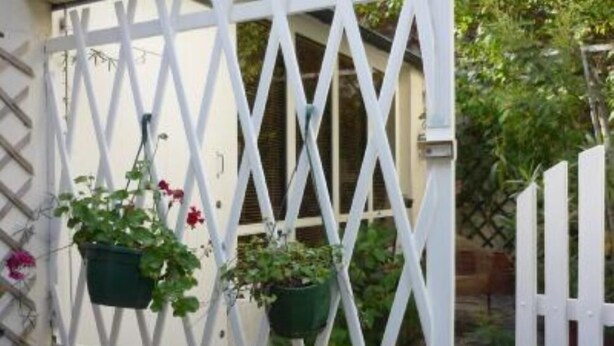 A white wooden fence with a lattice design, a window with hanging flower pots, and a view of greenery outside. - Paris 20 Pere Lachaise: VERY QUIET STUDIO WITH A TERRACE (Paris)