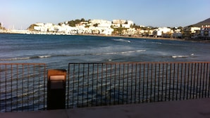 A coastal view with a metal railing in the foreground, a clear sky, and buildings along the shoreline.