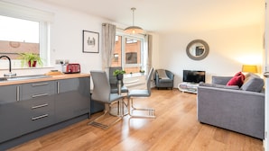 A modern kitchen with grey cabinets, a wooden countertop, and a red toaster. There is a dining area with a table and chairs, a grey sofa, and a TV. - York City Centre Luxurious Apartment with FREE ONSITE PARKING! (York)
