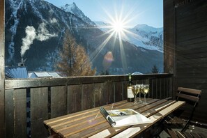Eine Holzterrasse mit Tisch, einer Weinflasche und Gläsern, mit Blick auf einen verschneiten Berg unter blauem Himmel.