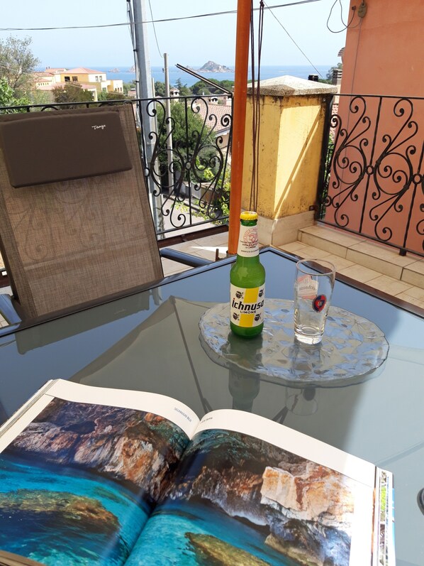 A glass table with a book, a bottle of Ichnusa lime, and a glass of water on a balcony with a view of the sea.