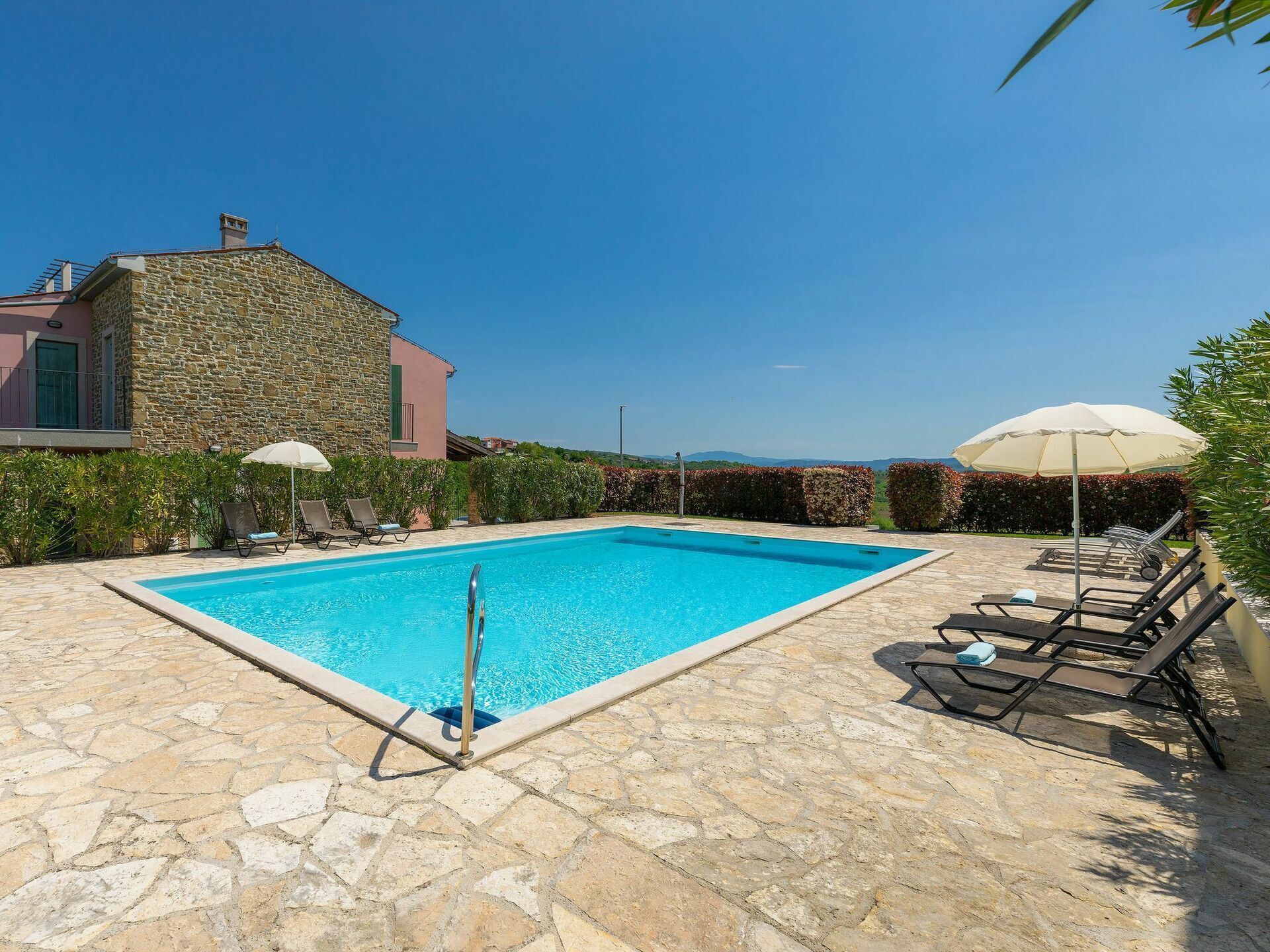 A rectangular swimming pool with a stone border, surrounded by lounge chairs and umbrellas, set against a backdrop of stone buildings and a clear sky.