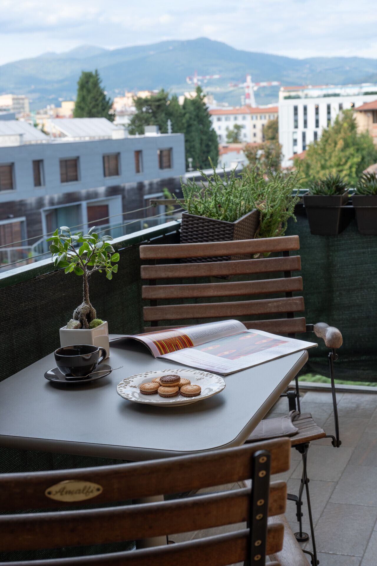 A table with a magazine, a cup, and a plate of cookies on a balcony overlooking a cityscape with buildings and mountains in the background.