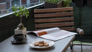 Ett bord med ett tidningsnummer, en kopp och en tallrik med kakor på en balkong med utsikt över en stadsbild, där byggnader och berg syns i bakgrunden.