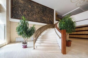 Una majestuosa escalera con barandilla de madera, flanqueada por plantas en macetas, que conduce a un techo alto con un gran mural.