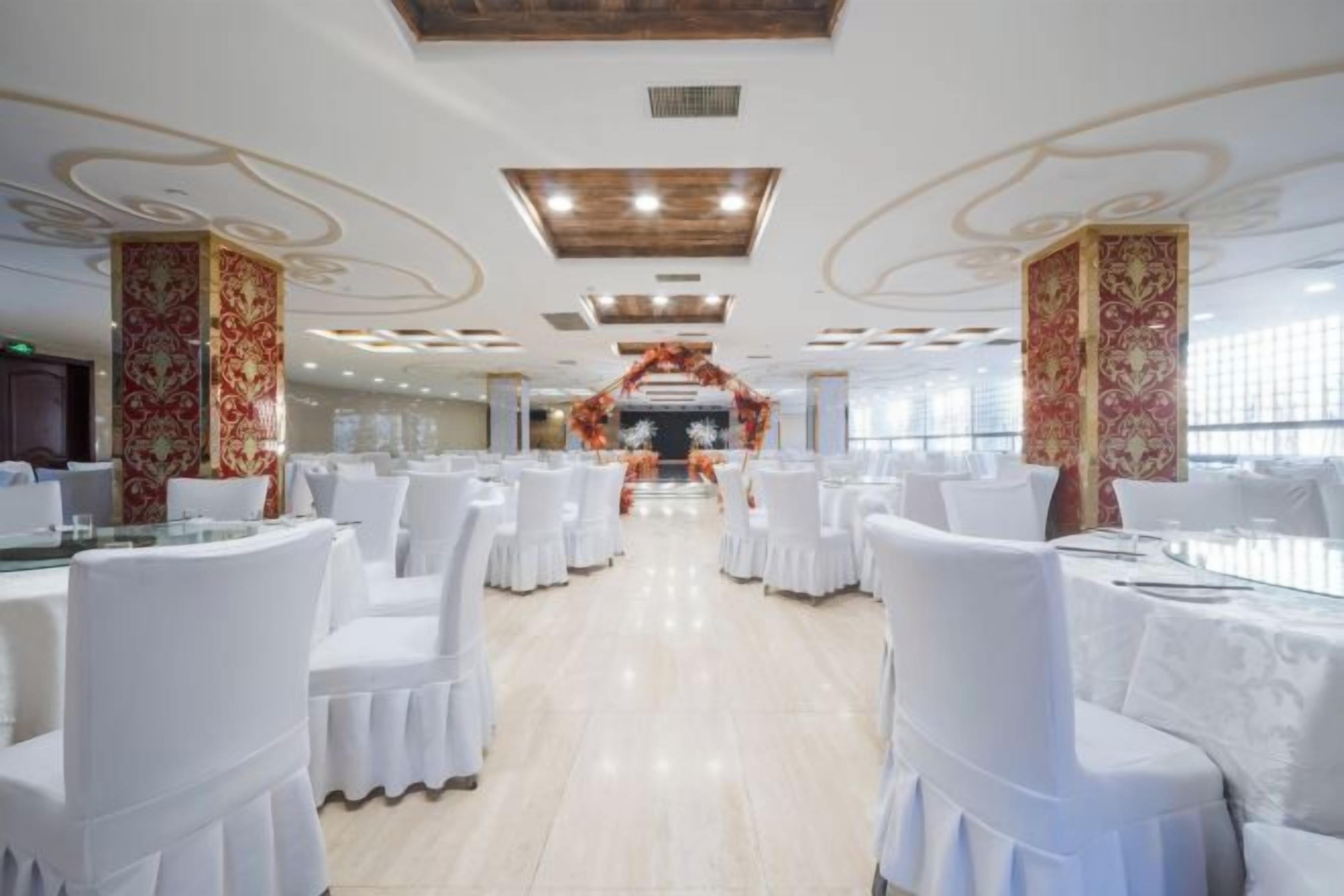 Ein Festsaal mit runden Tischen und Stühlen, einer Bühne mit Blumendekoration und einer hohen Decke mit eingelassener Beleuchtung.