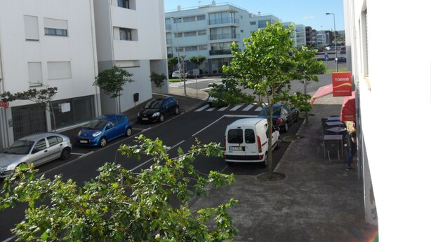 A parking area with several cars, a white van, and a blue car parked in front of a white building with multiple windows and balconies. - Chez Nônô - 4 bedroom, 2 toilet, in the city (Ponta Delgada)