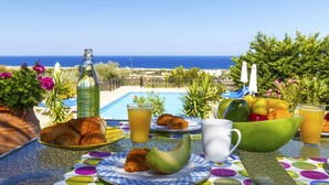 Ett frukostbord med croissanter, en melon och ett glas apelsinjuice, med utsikt över en pool och havet.