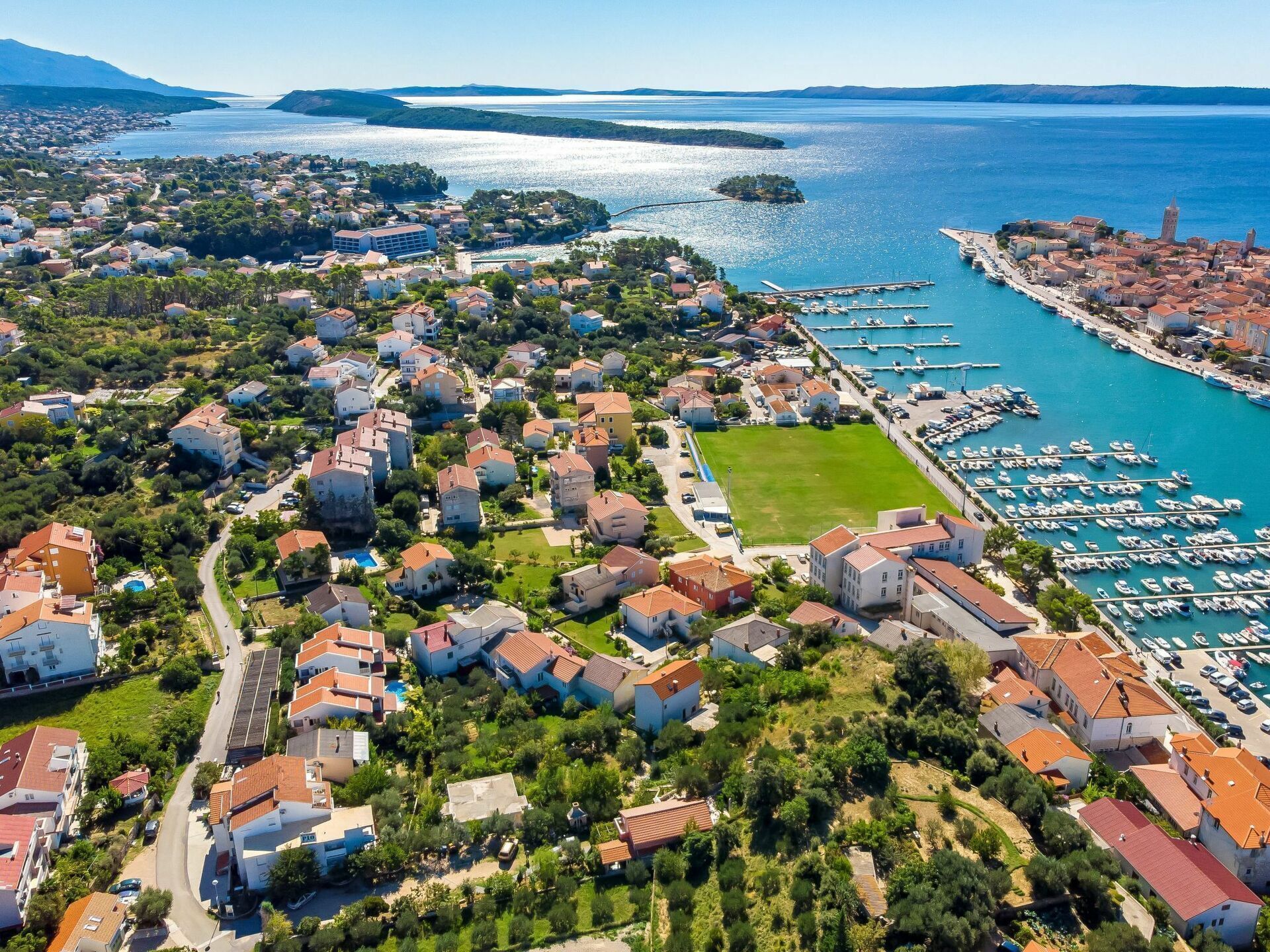 An aerial view of a coastal town with residential buildings, a marina, and a clear view of the sea.