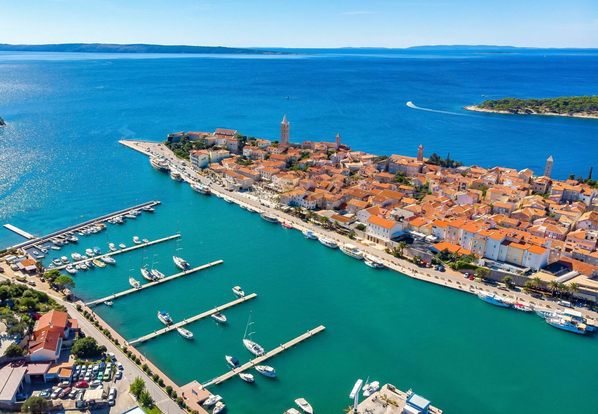 Vista aérea de un pueblo costero con un puerto deportivo, edificios con tejados rojos y un mar azul y despejado.