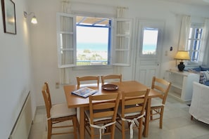 A dining area with a table and chairs, a view of the sea through open windows, and a lamp on a side table.