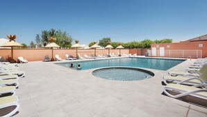Un área de piscina con sillas de descanso y sombrillas, cielo despejado y un edificio al fondo.