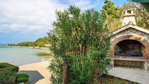 A stone-built outdoor kitchen with a brick oven, surrounded by greenery and a paved path leading to a beach.