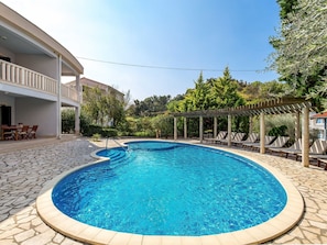 Uma piscina circular com deck de pedra, área de estar coberta com cadeiras e um edifício com varanda ao fundo.