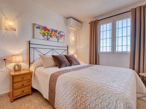 A bedroom with a bed, a nightstand, a lamp, a wooden dresser, a wall-mounted air conditioner, a window with curtains, and a colorful painting on the wall.