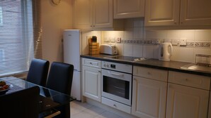 Una cocina moderna con armarios blancos, un refrigerador, un tostador y una tetera. Hay una mesa de comedor con sillas y una ventana con persianas.