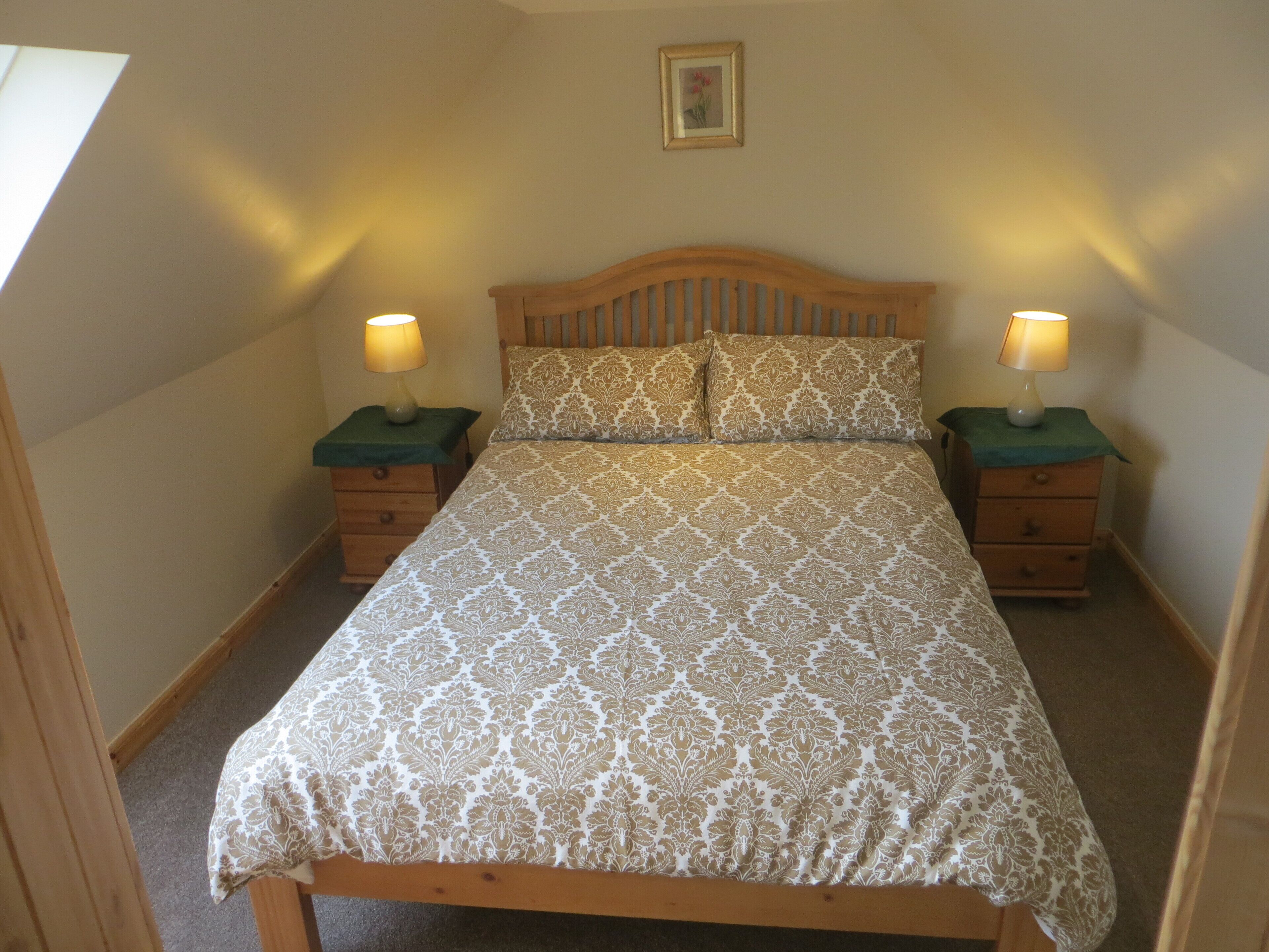 A bedroom with a wooden bed, two bedside tables with lamps, a framed picture on the wall, and a carpeted floor.