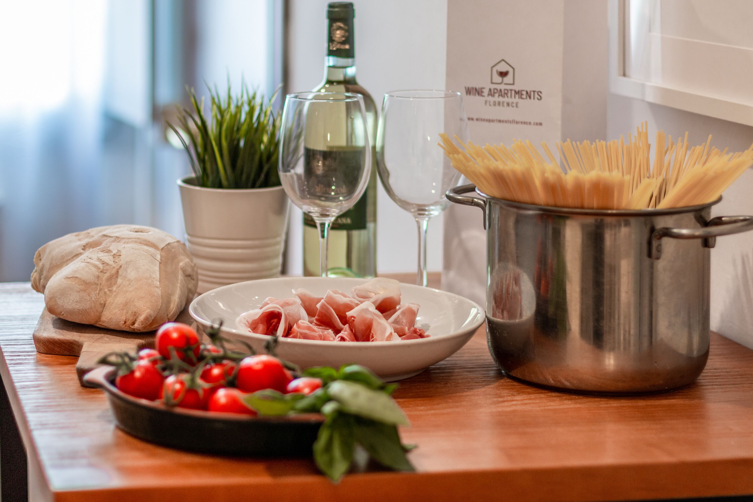 Eine Küchenarbeitsplatte mit einem Topf voller ungekochter Spaghetti, einer Schüssel mit geschnittenem Prosciutto, einem Brotlaib, einer Schüssel mit Cocktailtomaten und einer Weinflasche.