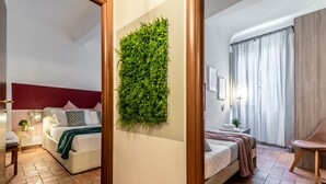A modern hotel room with a red accent wall, a green vertical garden, a bed with a white headboard, a bedside table with a lamp, and a chair.