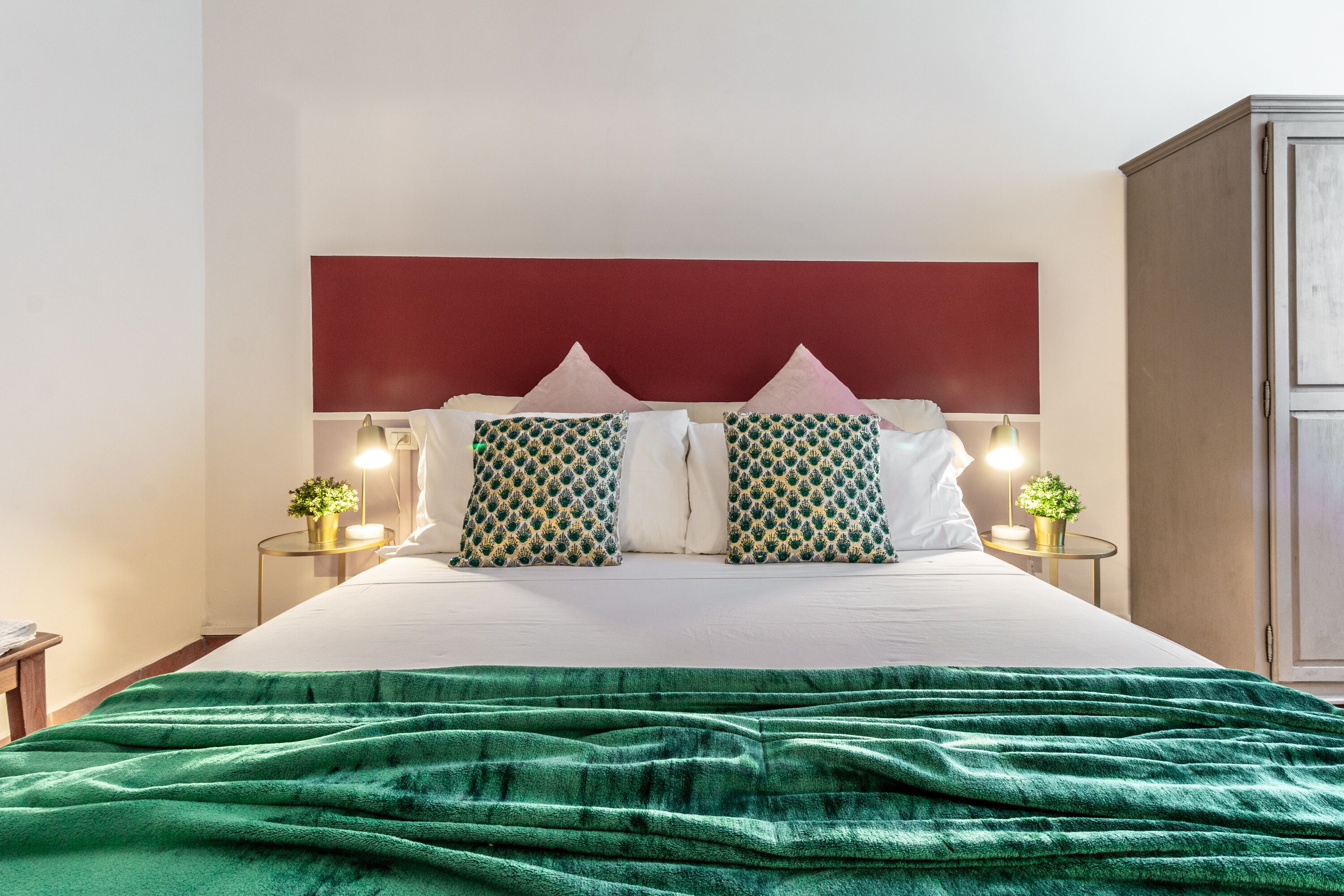 A neatly made bed with a green blanket, two patterned pillows, and a red headboard. There are bedside tables with lamps and small plants.