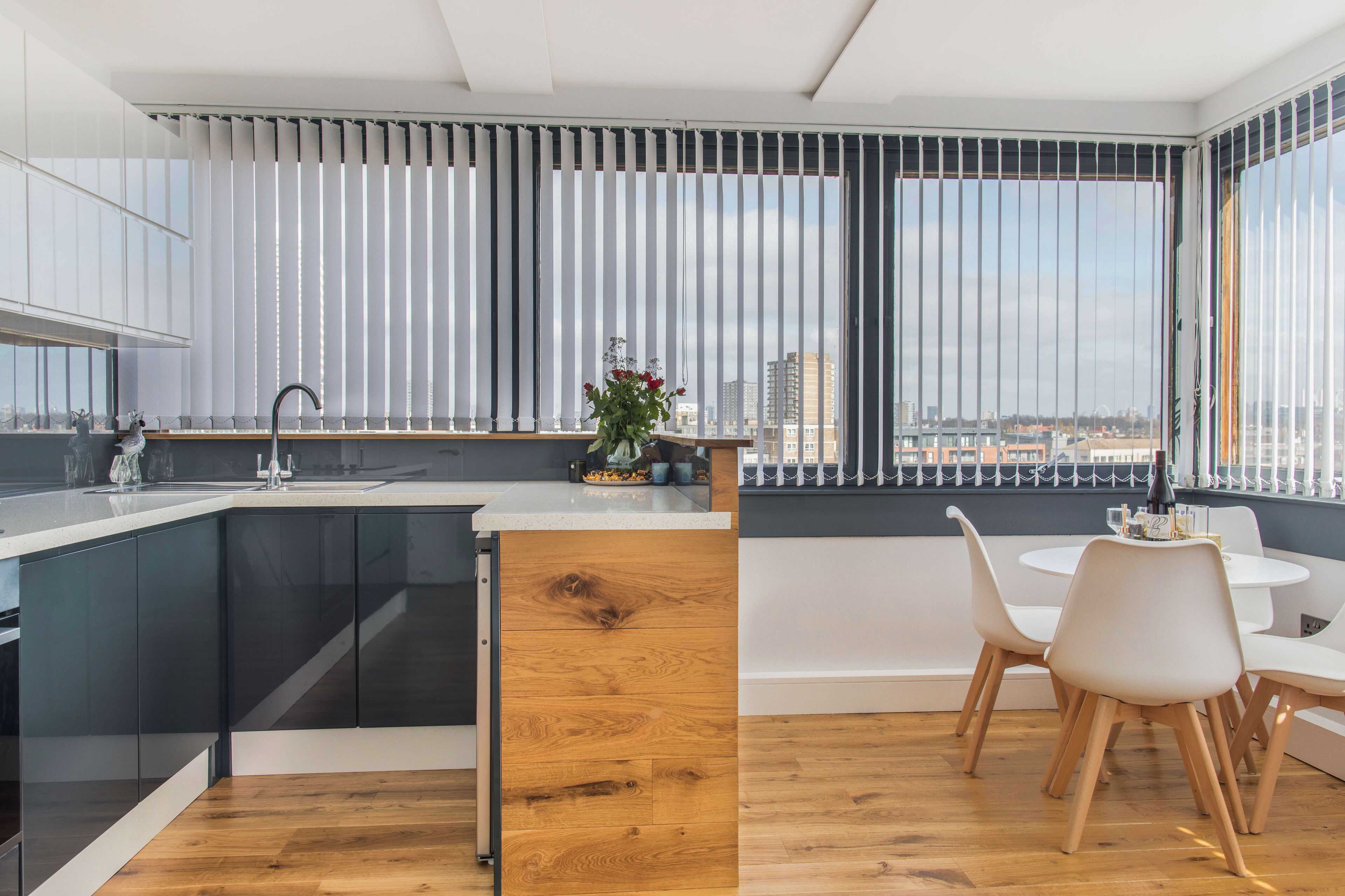 A modern kitchen with a wooden island, a dining area with white chairs, and large windows with blinds.