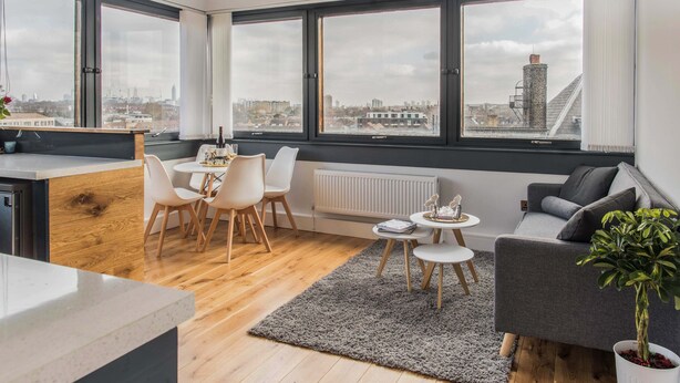 Un appartamento moderno con cucina, zona pranzo e soggiorno. Dispone di ampie finestre con vista sulla città, un divano grigio e una pianta in vaso.
