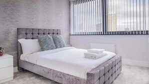 Una camera da letto moderna con un letto imbottito grigio, biancheria bianca e vista sulla città attraverso ampie finestre.