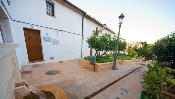 Un patio con un edificio blanco, una puerta de madera, una fuente de agua y plantas en macetas.