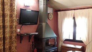 Una sala de estar con chimenea, televisor montado en la pared, una pequeña mesa de madera y un otoman con estampado.