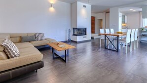 Uma sala de estar moderna com sofá, mesa de centro e televisão. Há uma área de jantar com mesa e cadeiras. O aposento possui uma lareira e uma cozinha ao fundo.