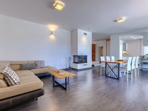 Ein modernes Wohnzimmer mit einer Couch, einem Couchtisch und einem Fernseher. Es gibt einen Essbereich mit Tisch und Stühlen. Im Hintergrund ist ein Kamin und eine Küche zu sehen.