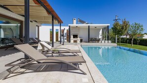 Uma área moderna com piscina ao ar livre, espreguiçadeiras, espaço para churrasco e céu azul.