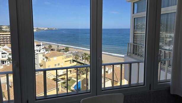 Blick aus einem Fenster auf einen Strand, ein Schwimmbecken und umliegende Gebäude.
