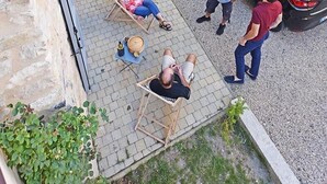 Edifício de pedra com porta de garagem, um carro preto estacionado na frente e pessoas sentadas e em pé numa área pavimentada.