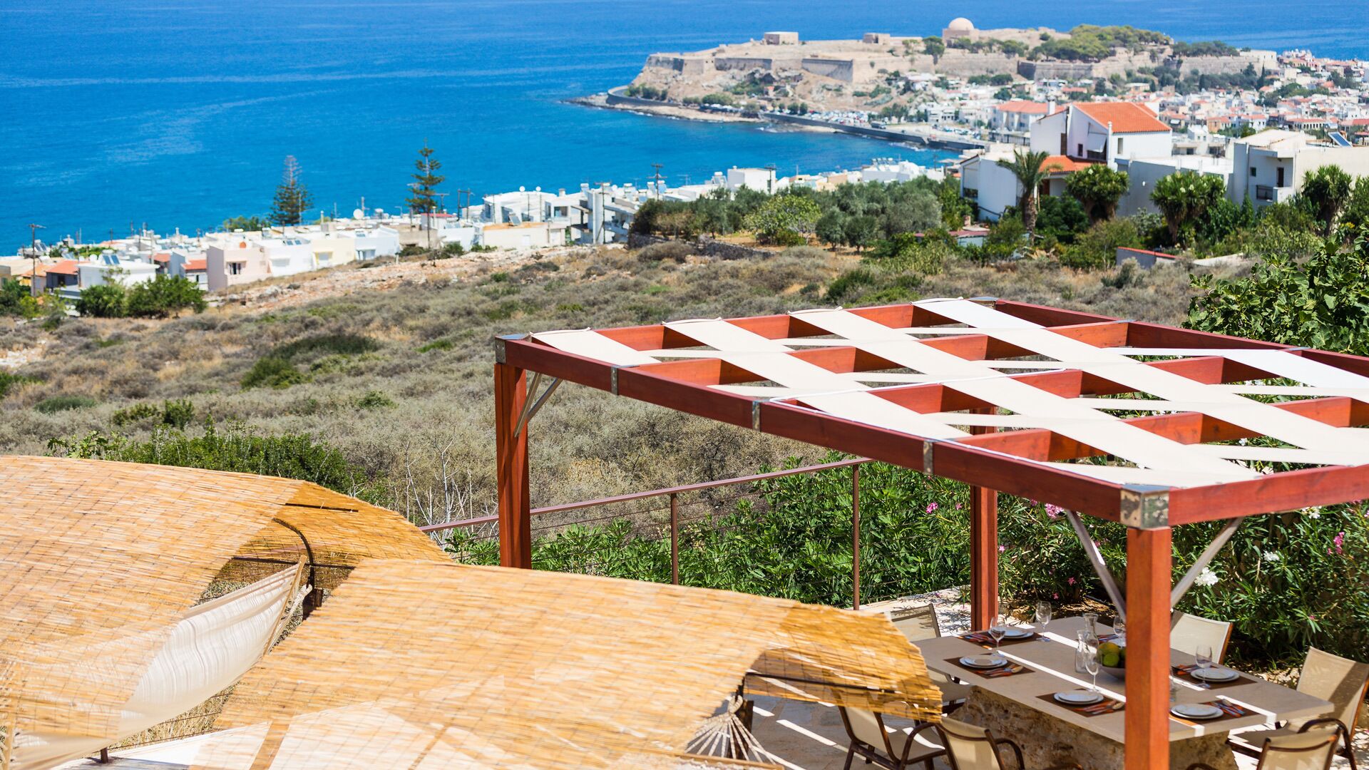 Une terrasse agrémentée d’une pergola en bois, une table dressée pour le repas, et une vue sur une ville côtière avec ses bâtiments et la mer.