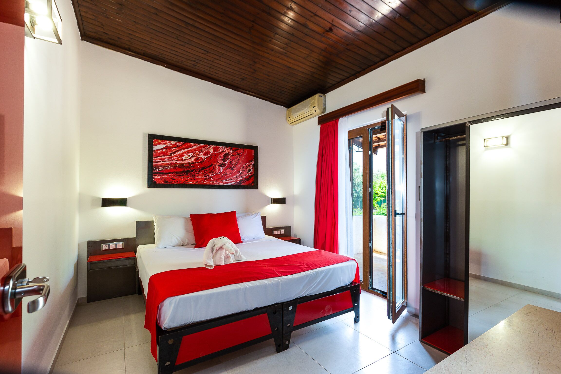A hotel room with a bed, a red and white bedspread, a red pillow, a nightstand, a wall-mounted artwork, and a sliding door leading to an outdoor area.